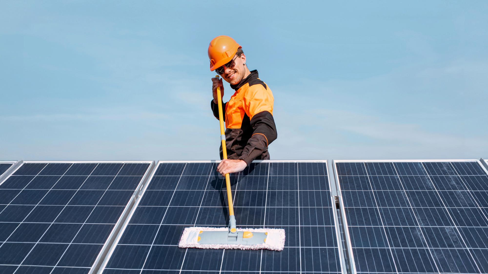 A worker in an orange hard hat and safety gear is cleaning solar panels with a long-handled brush, set against a clear blue sky.